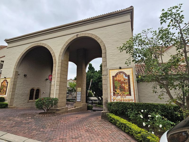 The main entrance to Via Positano featuring a prominent arched stone gateway, paved driveway, and decorative wall signage.
