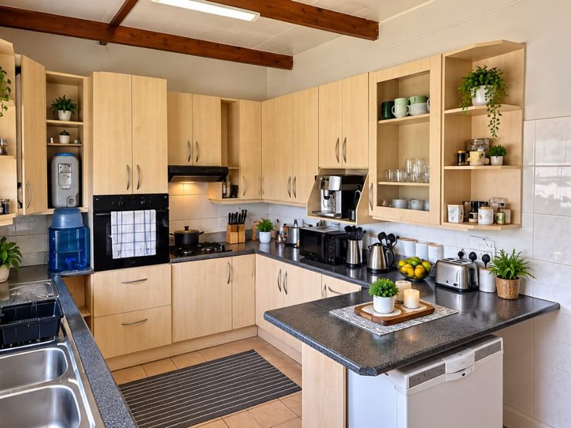 A functional kitchen featuring light wood cabinetry, dark laminate countertops, built-in oven and hob, and exposed timber ceiling beams with tiled flooring.