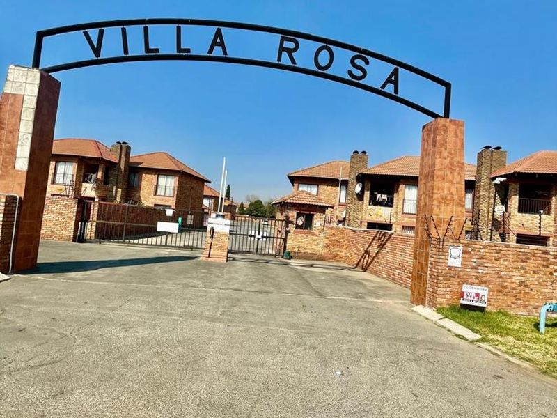 The entrance to Villa Rosa complex featuring a prominent metal signage arch, brick boundary walls, and a secure gated driveway.