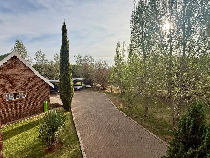 A view of the rear exterior featuring a brick building facade, a paved driveway leading to a carport, and established garden trees.
