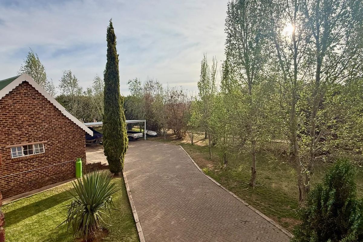 A view of the rear exterior featuring a brick building facade, a paved driveway leading to a carport, and established garden trees.