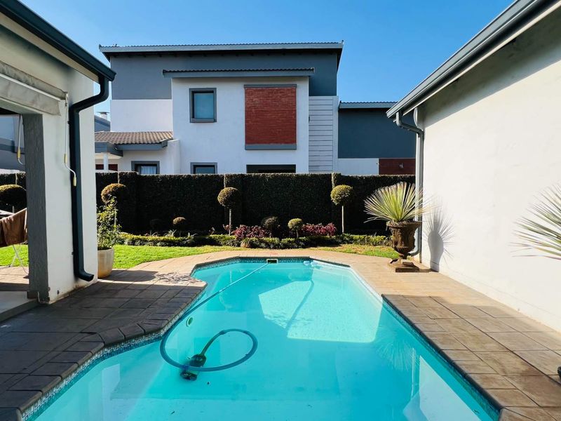 A well-maintained outdoor pool area featuring a tiled surround, a covered patio space, and a landscaped garden with a boundary wall.
