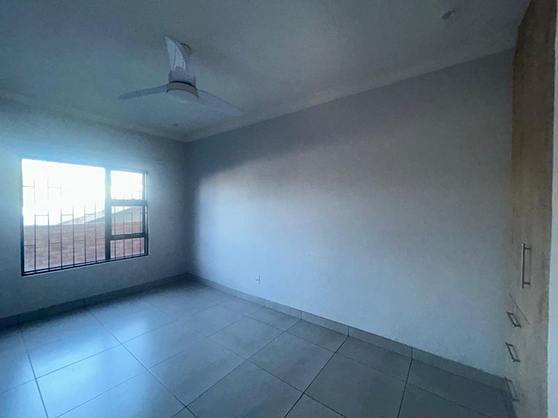 A bright, empty bedroom featuring tiled flooring, a ceiling fan, and built-in cupboards with security bars on the window.