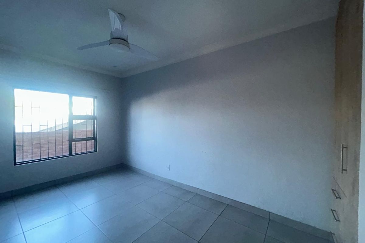 A bright, empty bedroom featuring tiled flooring, a ceiling fan, and built-in cupboards with security bars on the window.