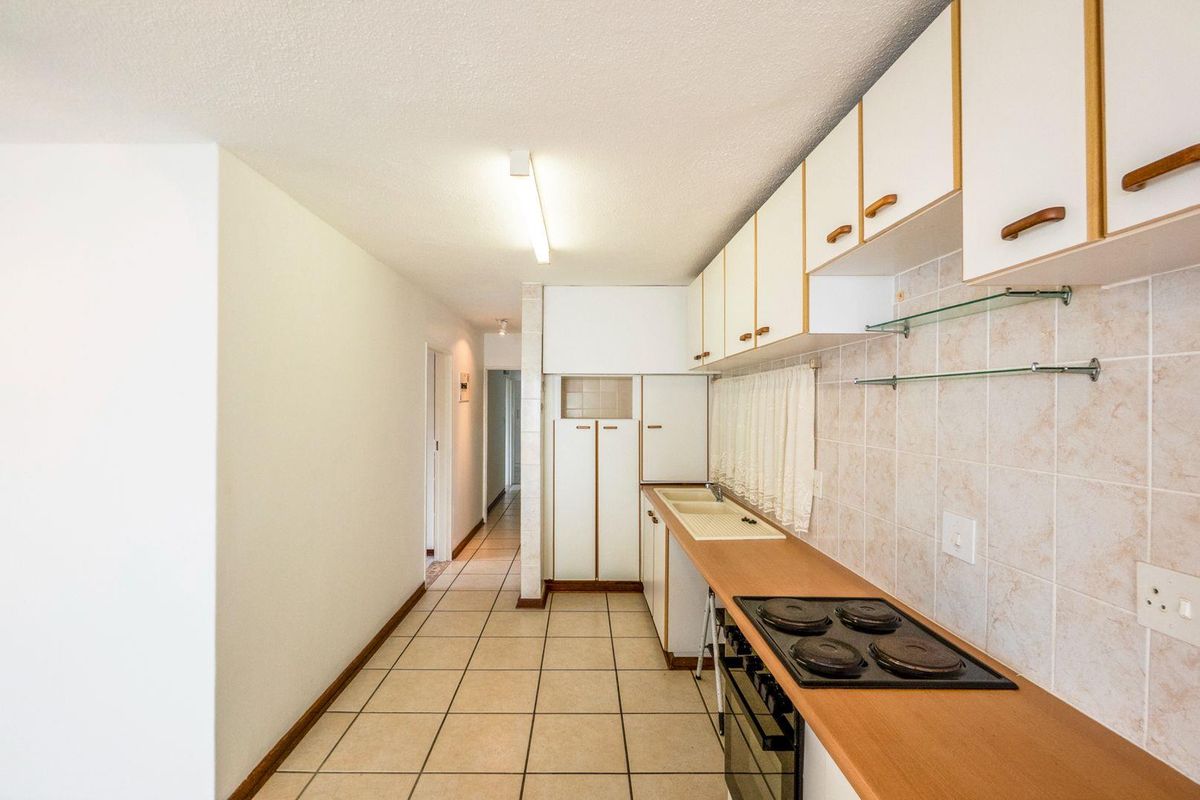 A functional galley-style kitchen featuring built-in cabinetry, a four-plate electric hob, integrated oven, and tiled flooring, leading into a hallway.