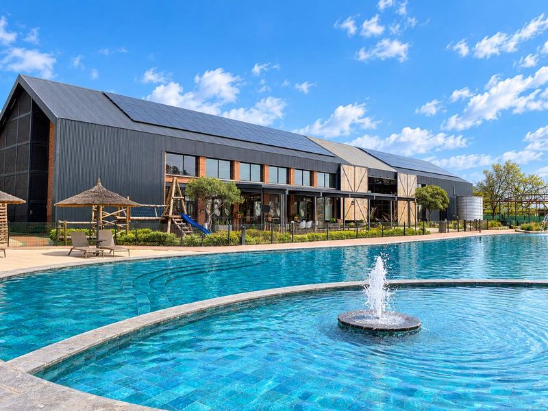 A modern apartment complex exterior featuring a large swimming pool with a central fountain, solar panels on the roof, and landscaped surroundings.