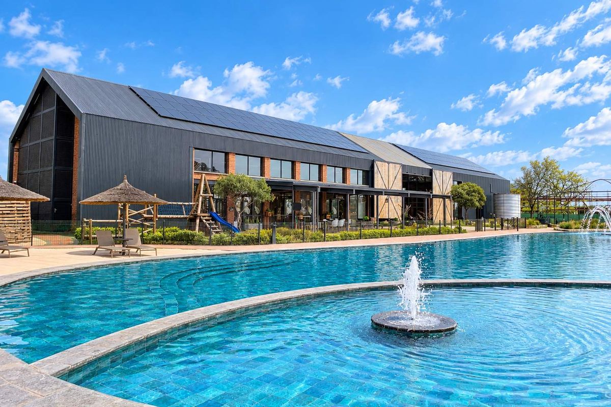 A modern apartment complex exterior featuring a large swimming pool with a central fountain, solar panels on the roof, and landscaped surroundings.