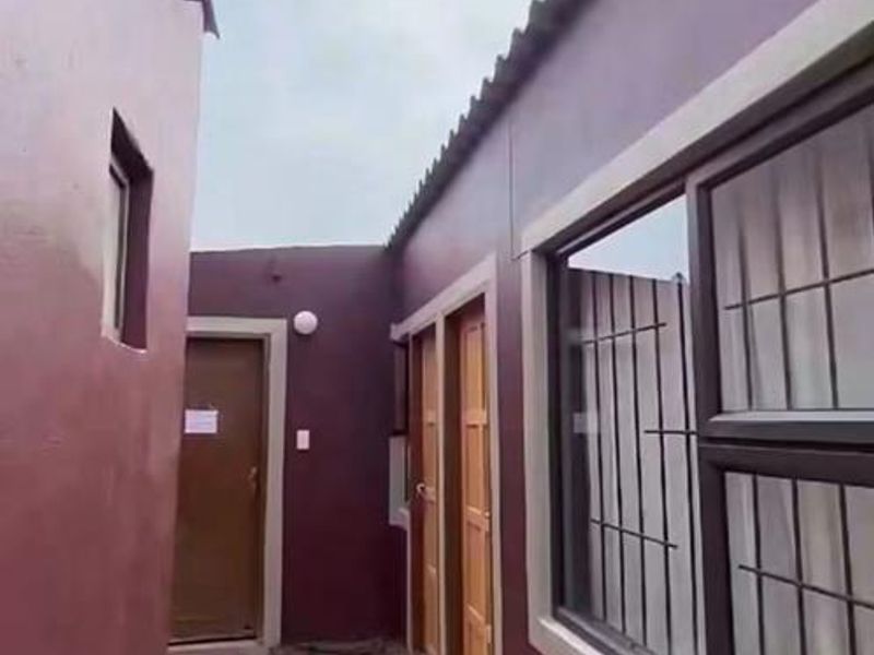 A narrow exterior side passage between two building sections featuring dark painted walls, aluminium windows with security bars, and an unfinished ground surface.