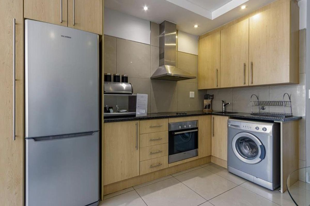 A compact, modern kitchen featuring light wood cabinetry, granite countertops, a stainless steel extractor fan, built-in oven, and integrated laundry appliance space.