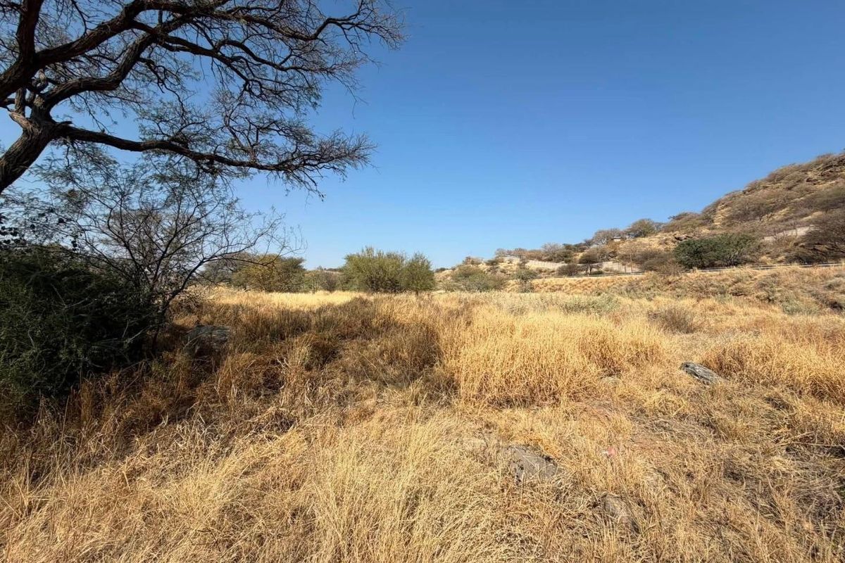 A view of a vacant commercial land plot featuring natural vegetation, dry grass, and rocky terrain under a clear blue sky.
