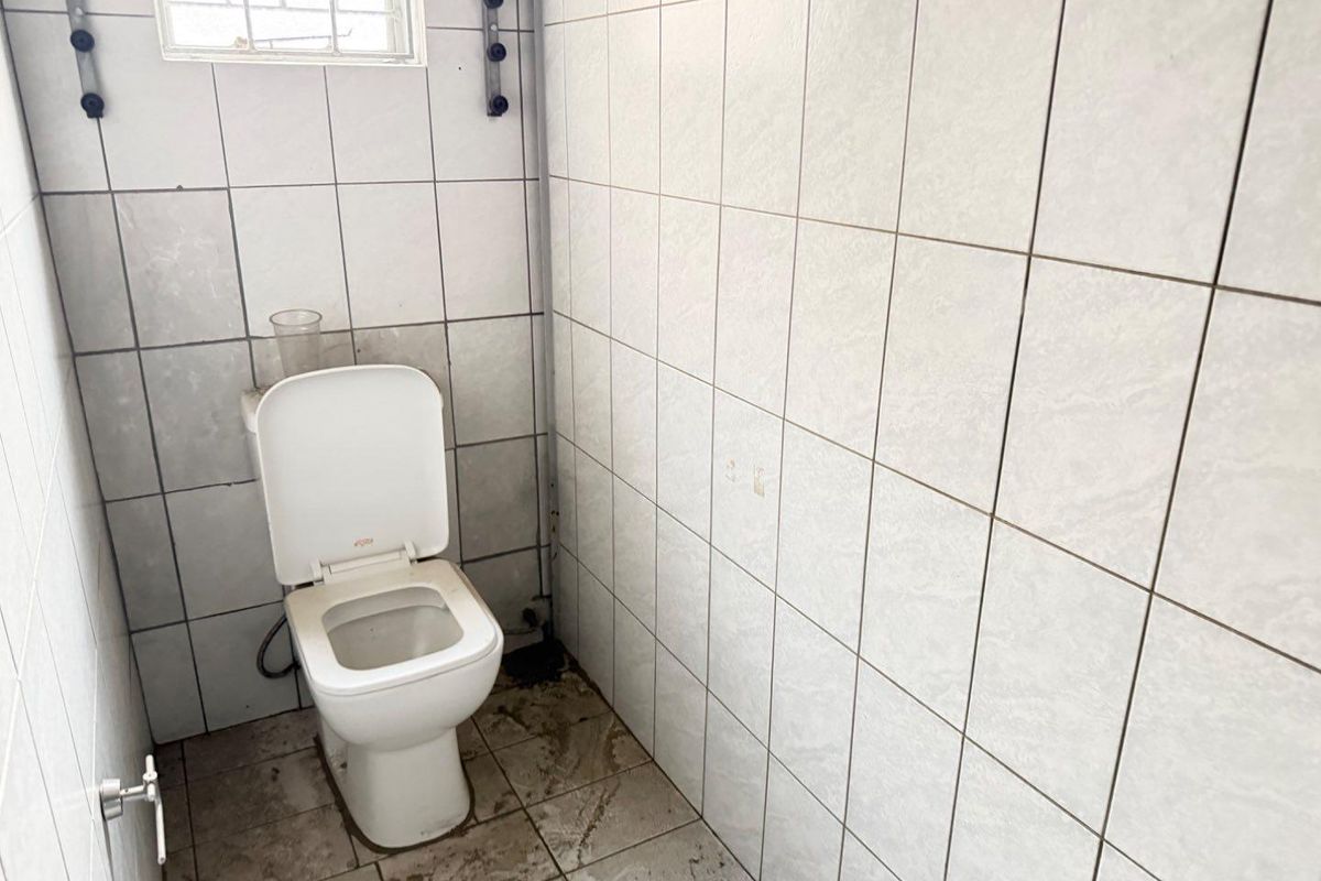 A small, functional bathroom featuring white wall tiles, a standard toilet, and a small window, requiring maintenance and cosmetic updates.