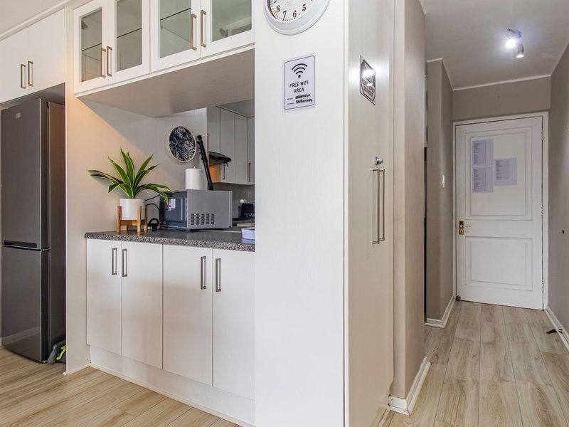 A compact, well-maintained kitchen area featuring modern white cabinetry, stone countertops, and light wood-look laminate flooring, leading into a hallway.