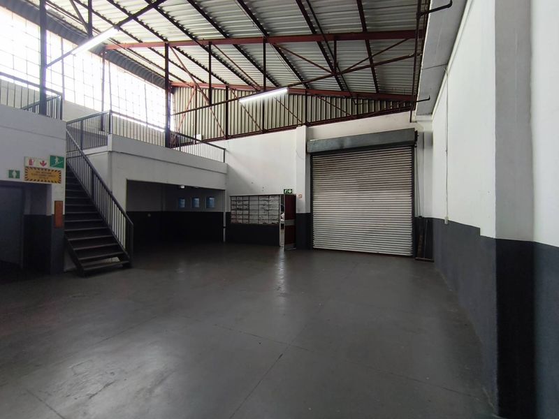 Spacious industrial warehouse unit featuring a high-clearance roof with exposed trusses, a roller shutter door, and a mezzanine level accessible via a steel staircase.