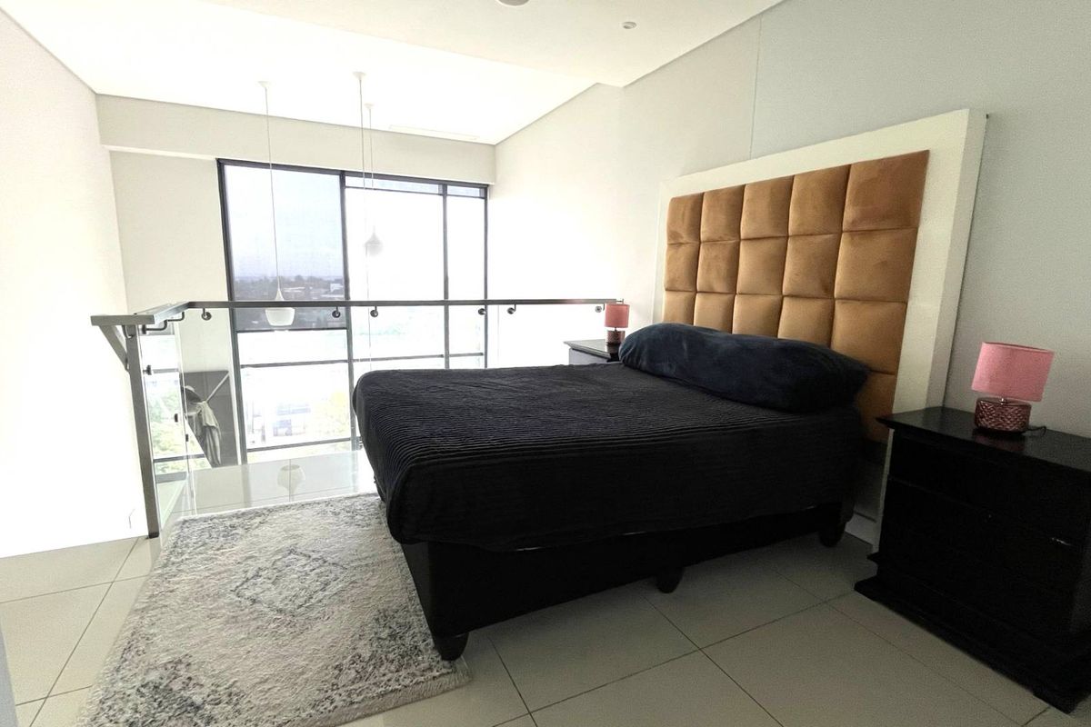 A modern mezzanine bedroom featuring tiled flooring, a glass balustrade overlooking the lower level, and large windows providing natural light.