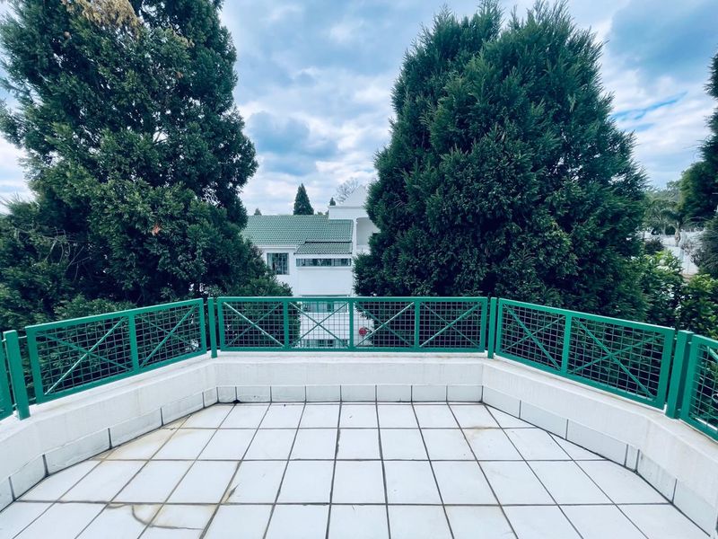 An open-air balcony featuring white tiled flooring and a green metal balustrade, offering a view of surrounding trees and neighboring buildings.