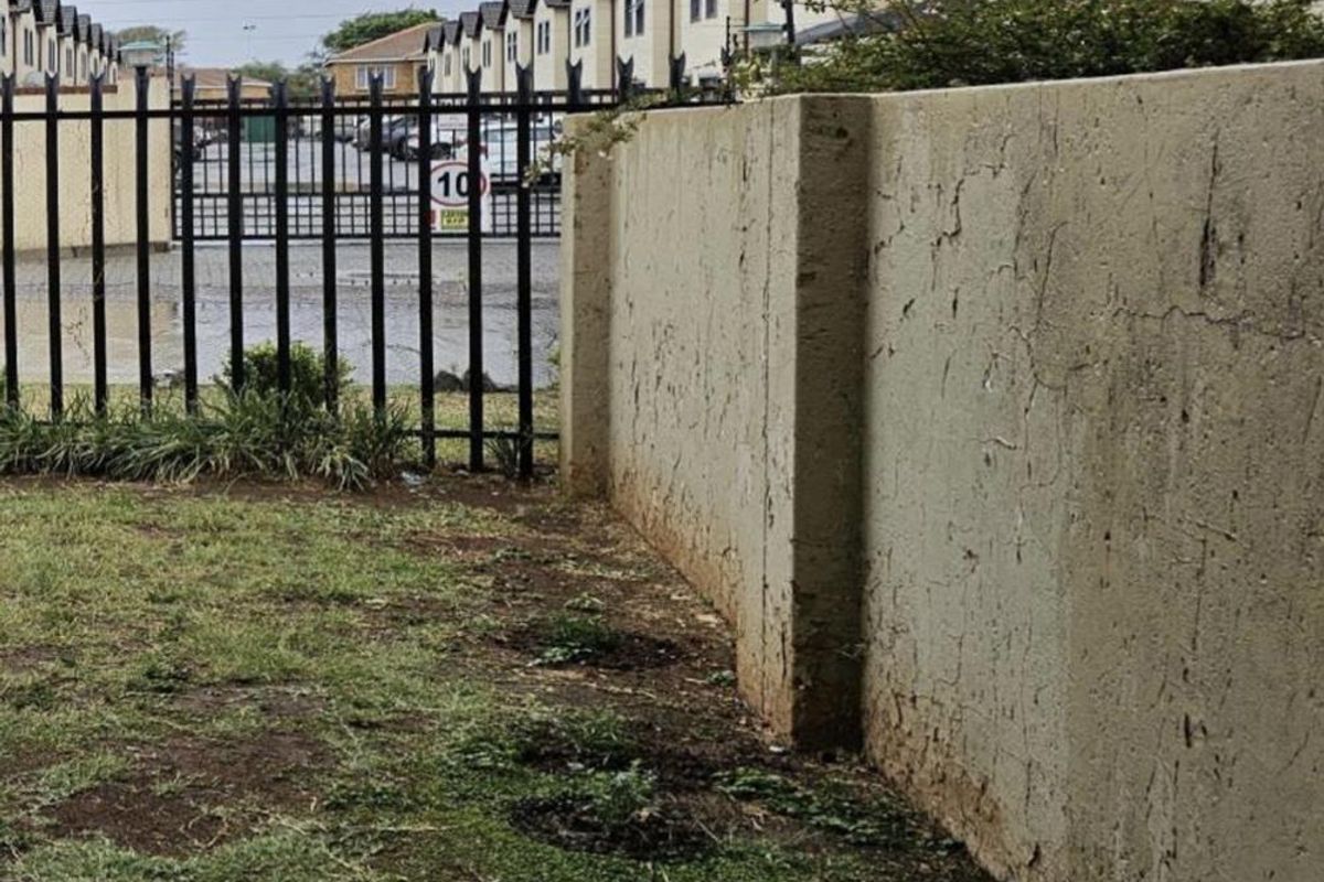 Exterior view of a residential complex boundary wall and metal security fencing with adjacent lawn area.