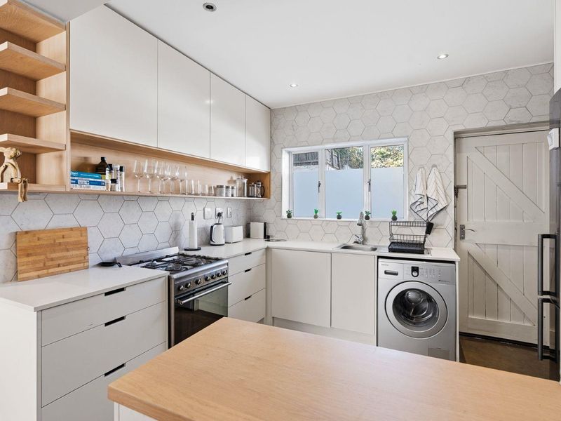 Modern kitchen featuring sleek white cabinetry, light wood accents, hexagonal tile backsplash, gas stove, and integrated laundry facilities.