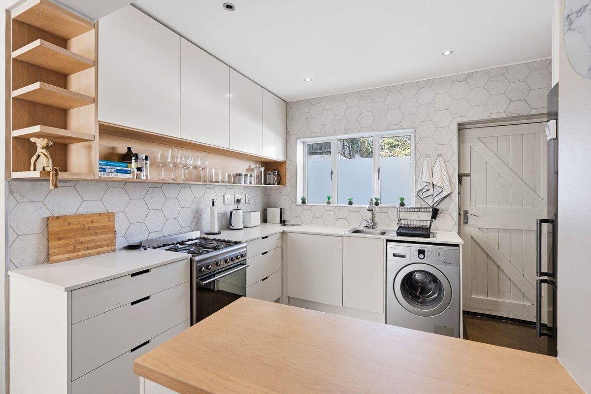 Modern kitchen featuring sleek white cabinetry, light wood accents, hexagonal tile backsplash, gas stove, and integrated laundry facilities.