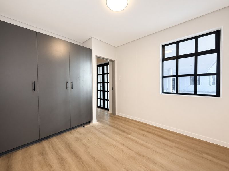 A bright, modern bedroom featuring light wood-look laminate flooring, sleek grey built-in cupboards, and a stylish black-framed window.