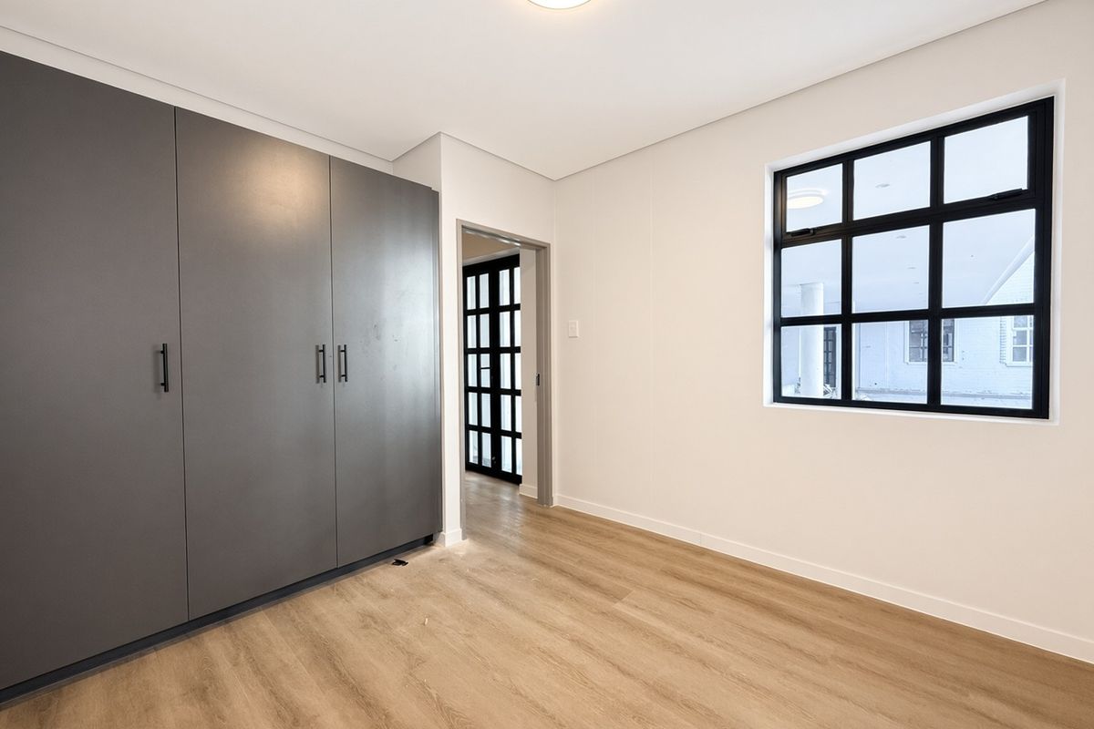 A bright, modern bedroom featuring light wood-look laminate flooring, sleek grey built-in cupboards, and a stylish black-framed window.