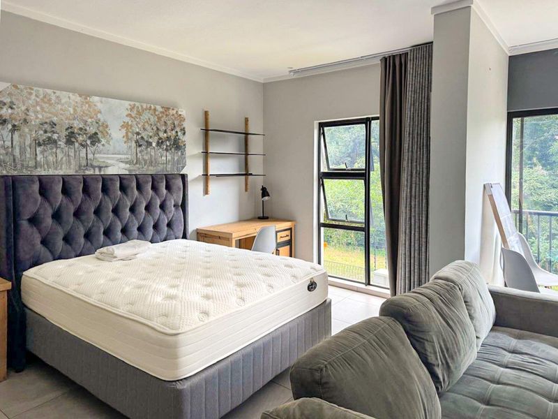 A well-lit bedroom featuring tiled flooring, neutral wall finishes, and large aluminium-framed windows providing natural light and garden views.