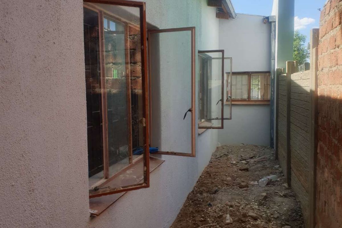 Side exterior view of the property showing unfinished ground area, boundary wall, and multiple open steel-framed windows along the building facade.
