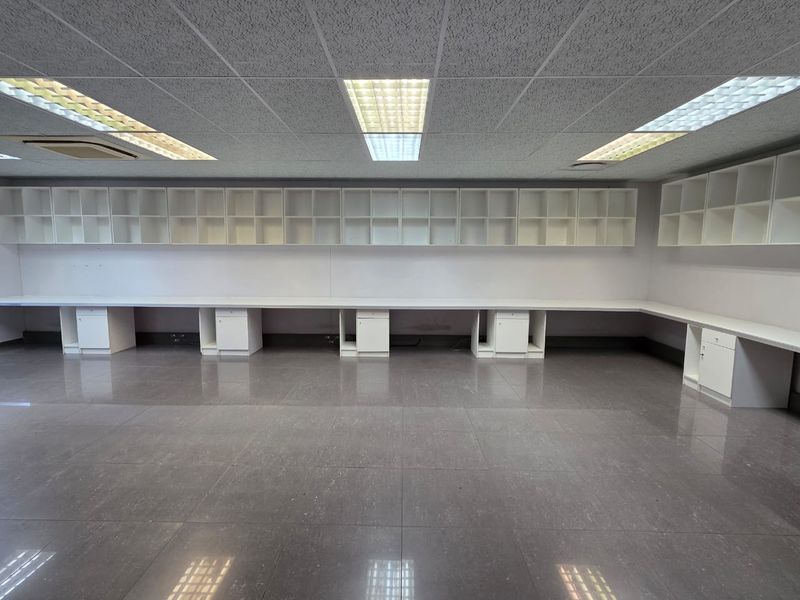 A spacious, empty office area featuring extensive built-in wall cabinetry, integrated desk workstations, suspended ceiling tiles, and durable tiled flooring.