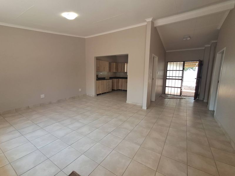 Spacious open-plan living area featuring neutral tiled flooring and a functional kitchen with built-in cabinetry, leading to an exterior security gate.
