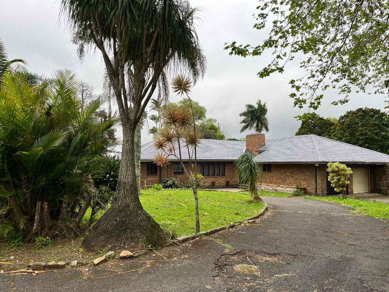 Front view of a single-story brick house featuring a tiled roof, brick chimney, attached garage, and a mature landscaped garden with an asphalt driveway.