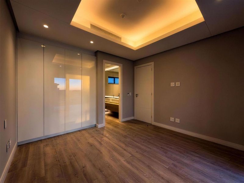Modern bedroom featuring high-gloss built-in wardrobes, warm cove lighting, and wood-look laminate flooring, with an adjoining en-suite bathroom.