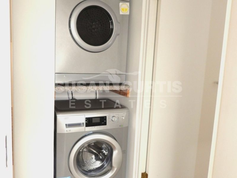 A compact laundry nook featuring a stacked washing machine and tumble dryer unit with a stone shelf, set within a clean, modern apartment space.