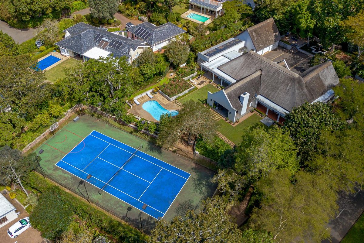 Aerial view of a large residential property featuring a private tennis court, swimming pool, landscaped gardens, and a main house with a thatched roof.