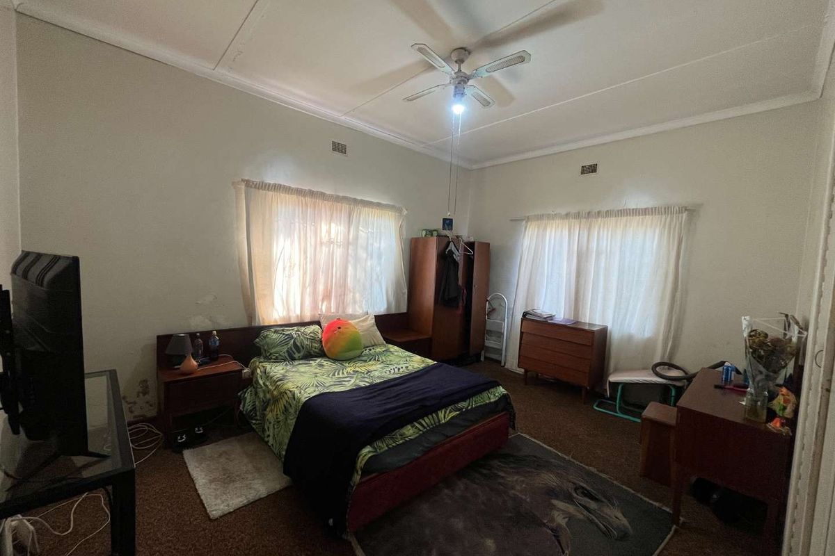 Spacious bedroom featuring a ceiling fan, built-in headboard, and dual windows. The room requires cosmetic updates including wall repairs and new flooring.