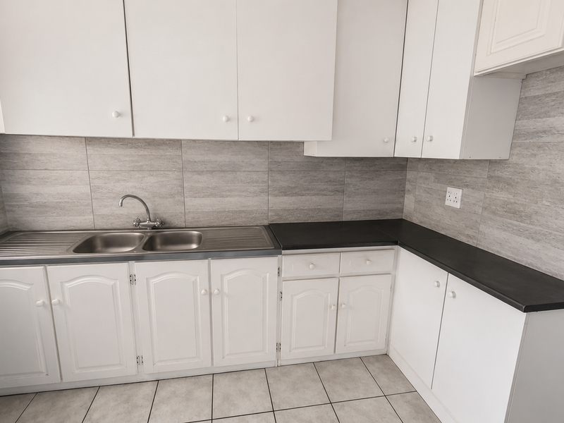 A clean, functional kitchen featuring white cabinetry, dark laminate countertops, a double stainless steel sink, and neutral tiled backsplash and flooring.