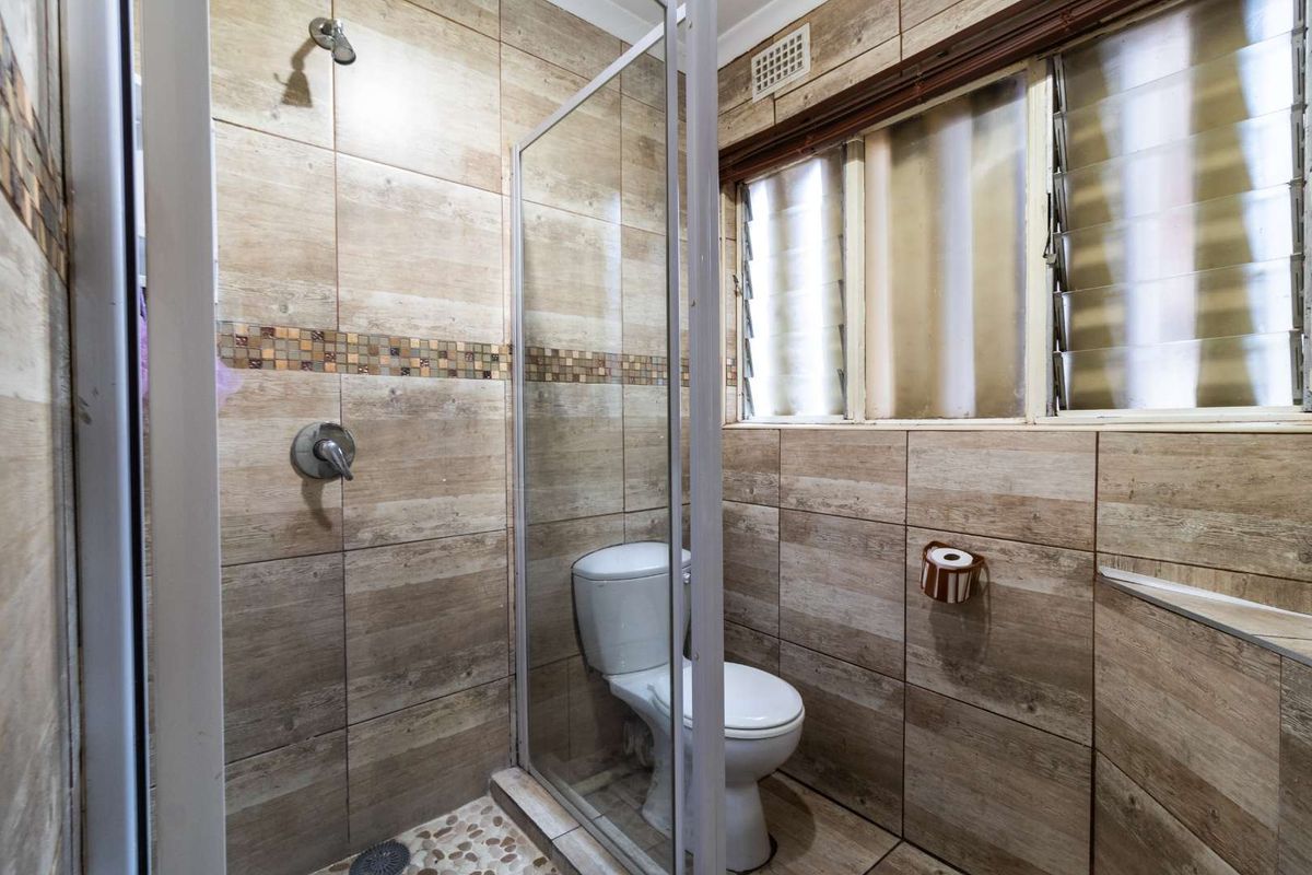 A functional bathroom featuring a glass-enclosed shower, toilet, and wood-look ceramic wall tiling with louvered windows for ventilation.