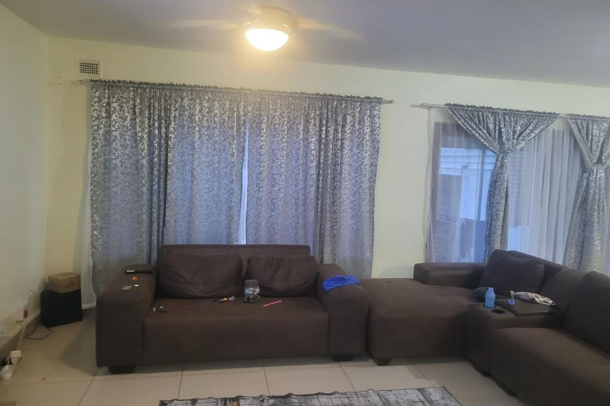 A living room featuring tiled flooring, a ceiling fan, and large windows with curtains. The space shows signs of wear on the floor.
