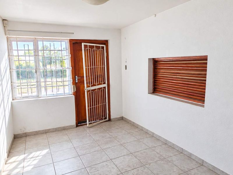 A bright, empty office space featuring tiled flooring, a secure entrance door, and natural light through a window with security bars.