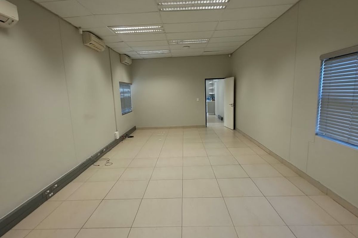 A spacious, empty office area featuring tiled flooring, suspended ceilings with recessed lighting, and multiple wall-mounted air conditioning units.