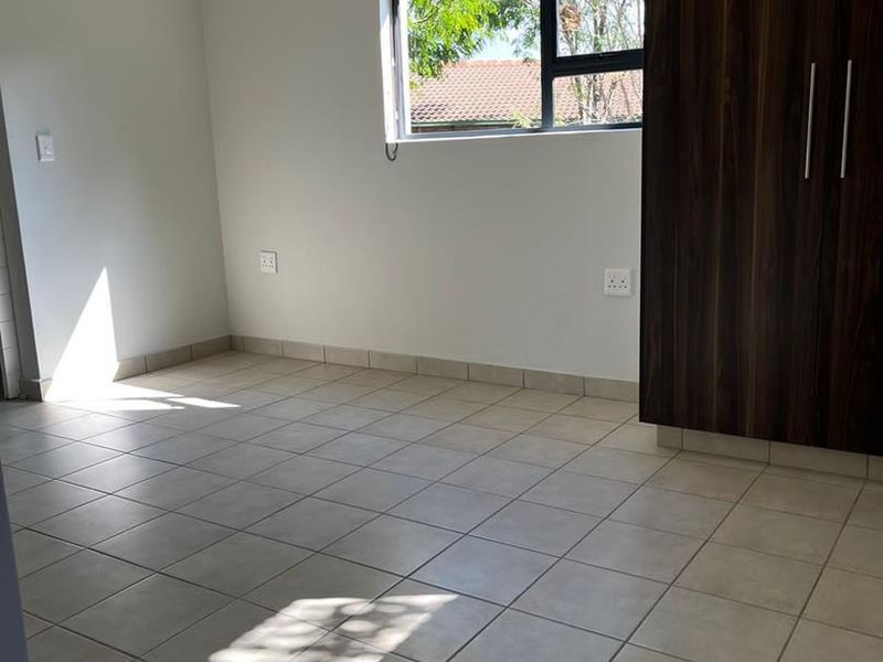 A bright, empty bedroom featuring neutral tiled flooring, a built-in dark wood wardrobe, and a window with natural light.