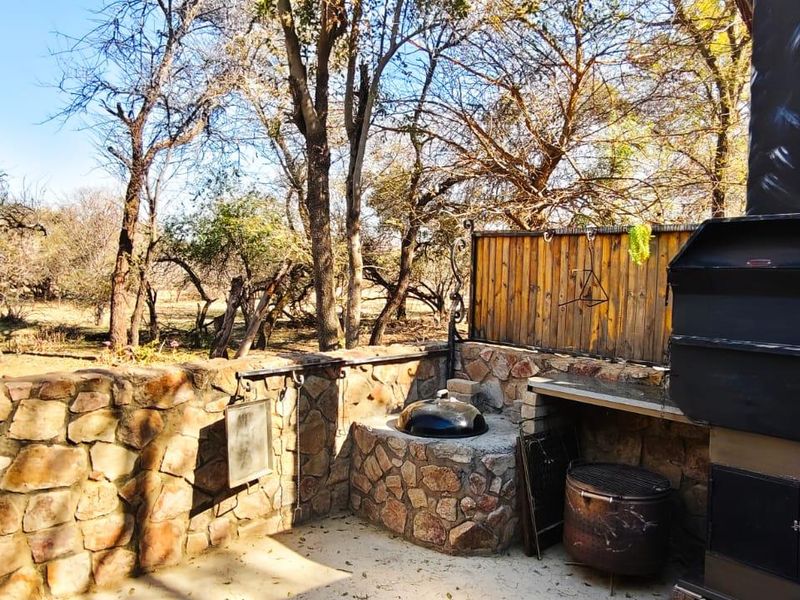Outdoor entertainment area featuring a built-in braai, stone masonry walls, and a concrete prep surface, set against a natural wooded backdrop.