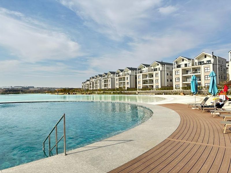 A well-maintained communal swimming pool area featuring a large pool with a concrete surround and composite decking, set against a modern apartment complex.