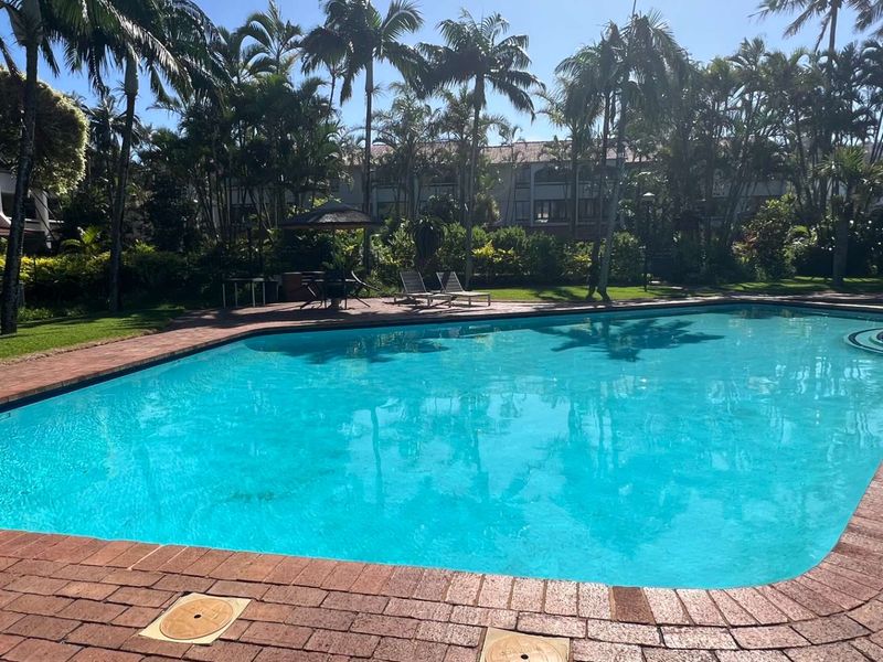 A well-maintained communal swimming pool area surrounded by lush tropical landscaping and mature palm trees, featuring brick paving and clear water.
