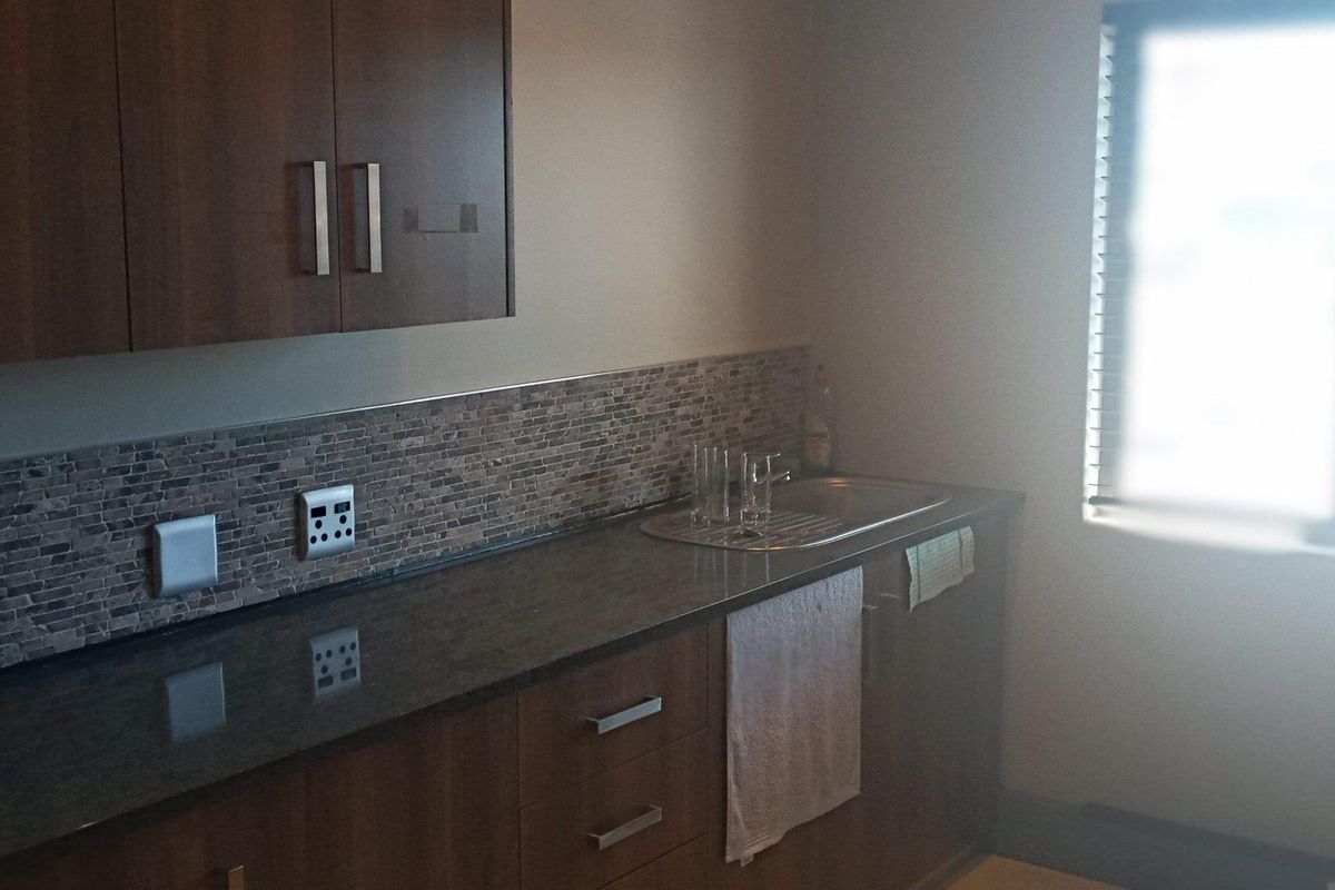 A functional office kitchenette featuring dark wood cabinetry, stone-look backsplash, granite-style countertop, and a stainless steel sink.