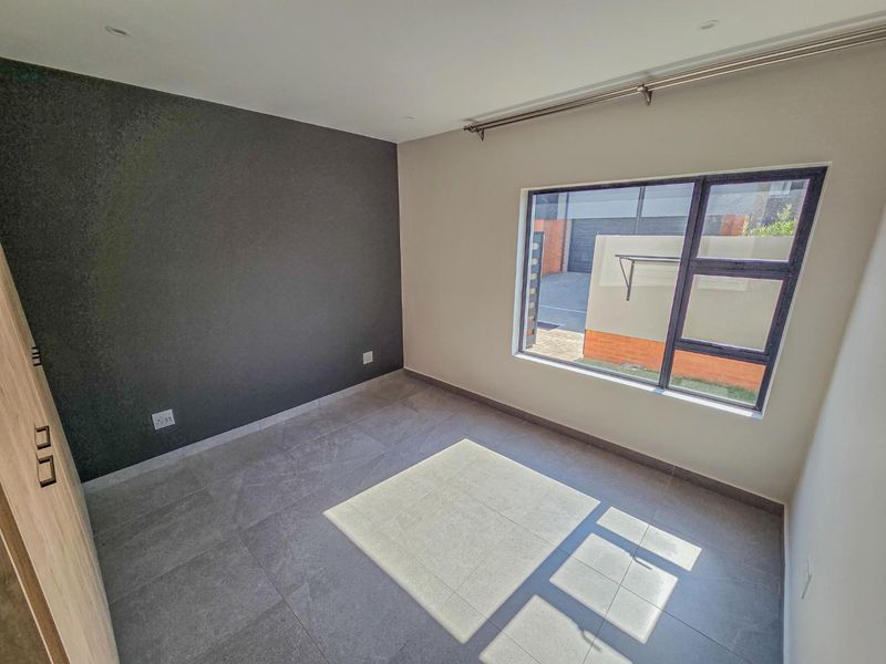 A bright, empty bedroom featuring modern grey floor tiles, a dark accent wall, built-in cupboards, and large aluminium-framed windows.