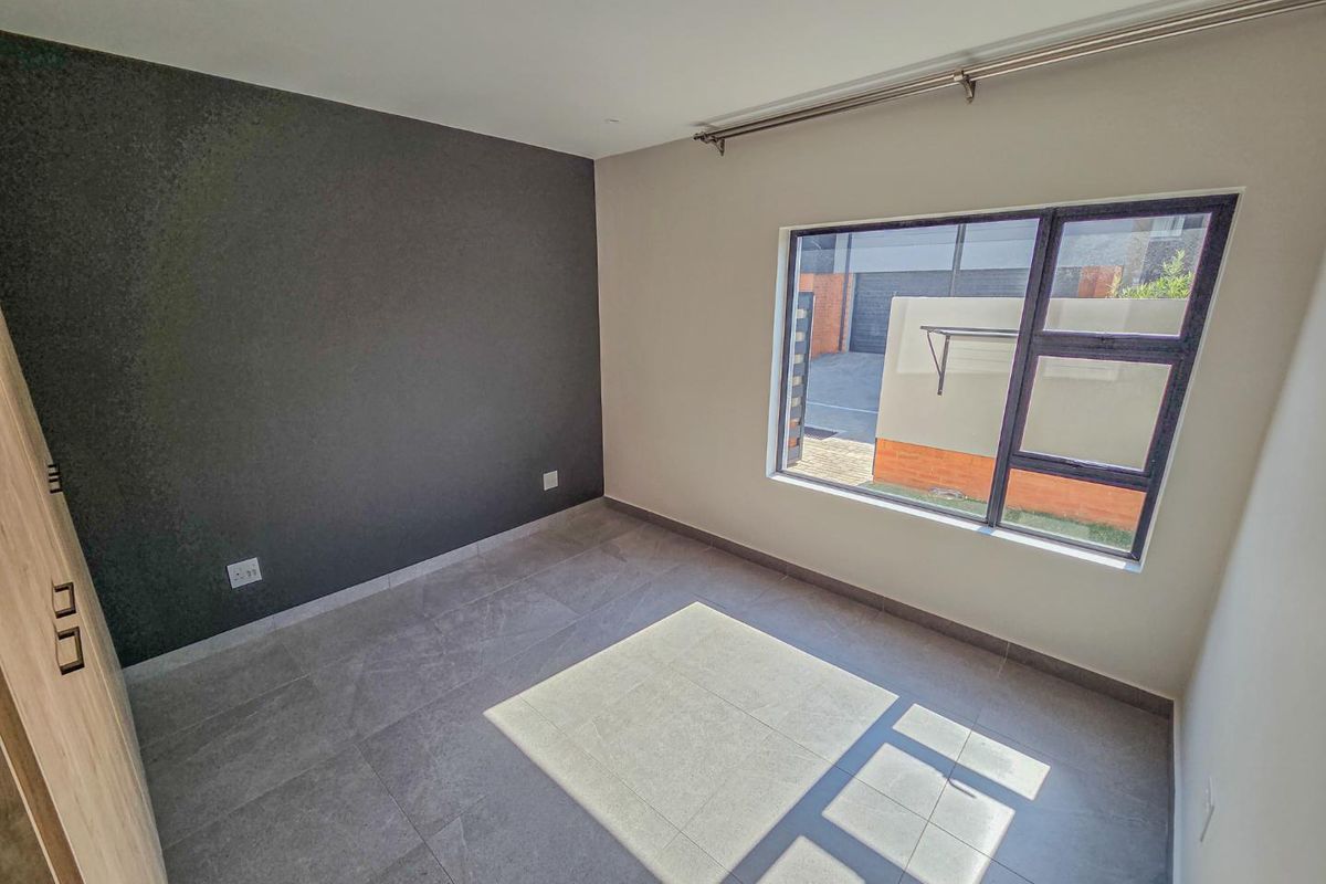 A bright, empty bedroom featuring modern grey floor tiles, a dark accent wall, built-in cupboards, and large aluminium-framed windows.