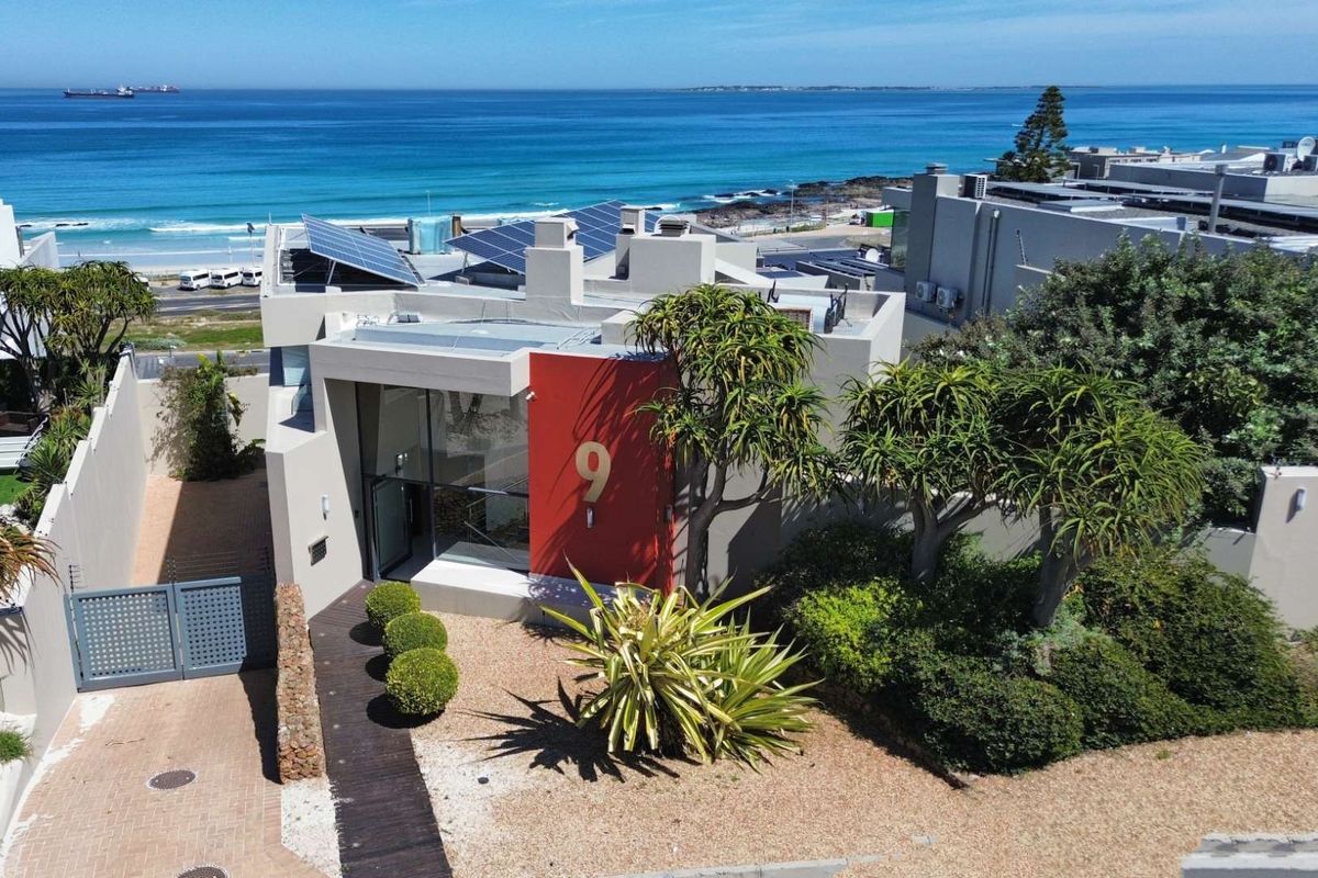 Aerial view of a modern coastal property featuring solar panels, a landscaped garden, and a paved driveway, situated near the ocean.