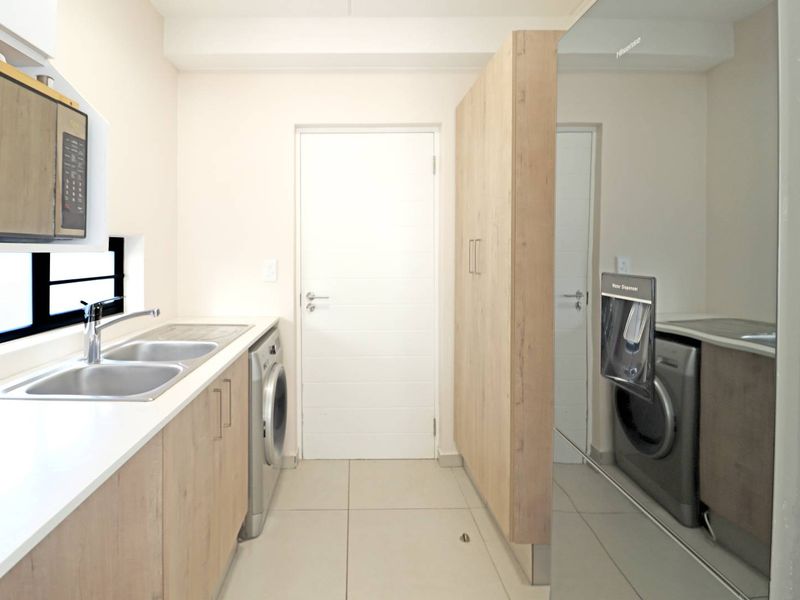 A modern, functional laundry room featuring light wood-finish cabinetry, a double sink with mixer tap, and tiled flooring.
