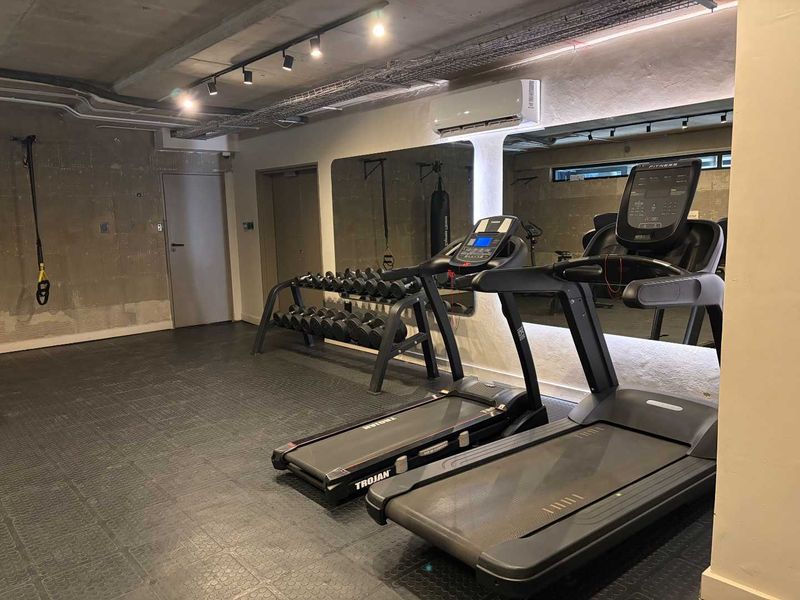 A modern, industrial-style gym featuring exposed concrete ceilings, rubber flooring, wall-mounted mirrors, and integrated air conditioning.