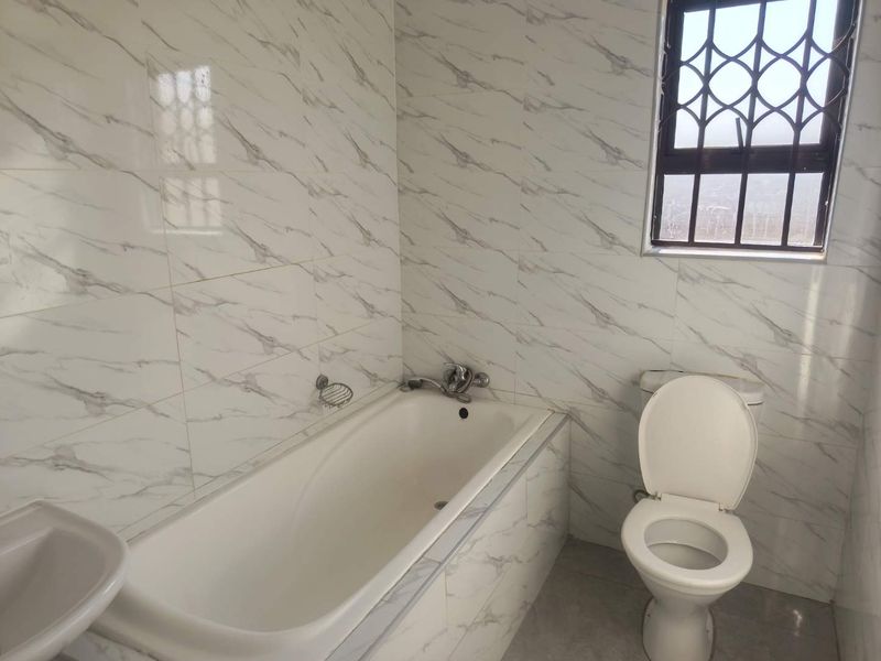 A clean, functional bathroom featuring marble-look wall tiles, a built-in bathtub, a standard toilet, and a washbasin with a security-grille window.
