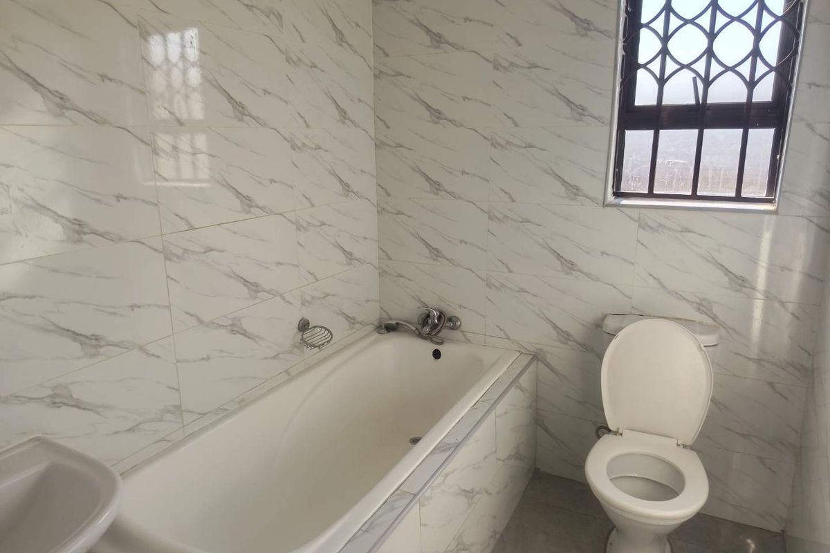 A clean, functional bathroom featuring marble-look wall tiles, a built-in bathtub, a standard toilet, and a washbasin with a security-grille window.